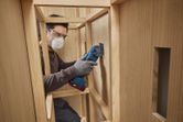 A person wearing safety equipment uses a multifunction tool to cut wood inside a cabinet.