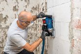 A person wearing safety equipment uses a wall chaser on a concrete wall.