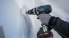Person wearing safety equipment drills a hole into a wall with a cordless power tool.