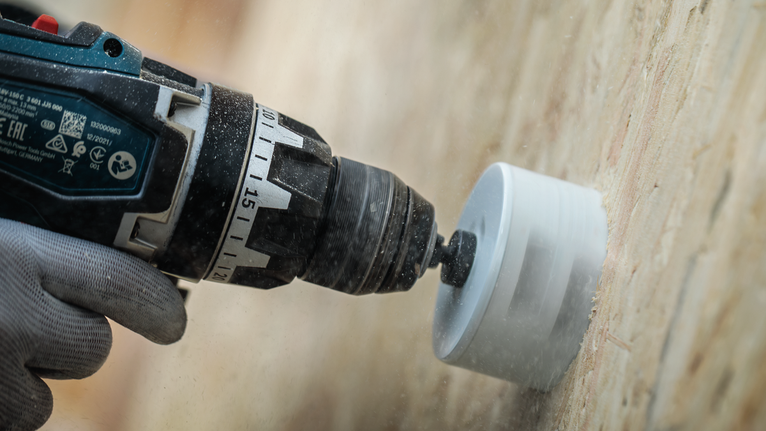 Person wearing safety equipment drills a hole into wood with a hole saw attachment.