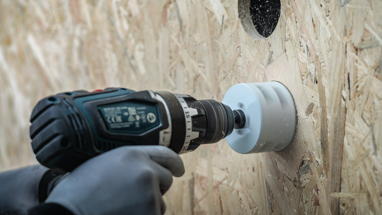 Person wearing safety equipment drilling a circular hole in a wooden board.