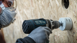 Person wearing safety equipment uses a drill with a hole saw on a wooden panel.