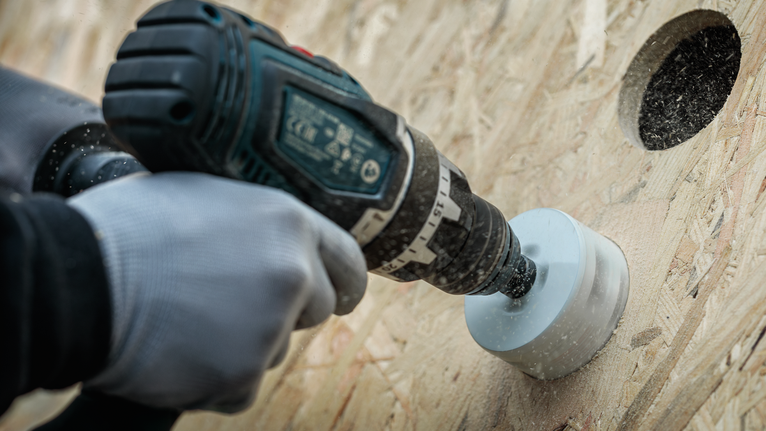 Person wearing safety equipment drills a circular hole in plywood with a hole saw.