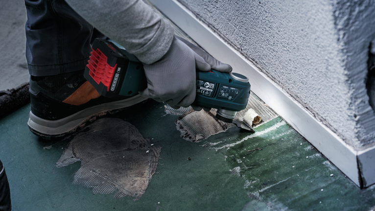 Person wearing safety equipment cuts flooring adhesive near a wall with a power tool.
