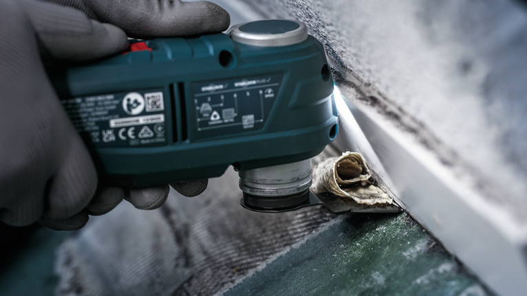 Person wearing safety equipment removes old sealant from a wall using a multi-tool.