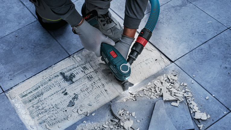 Person wearing safety equipment removes tile adhesive with a power tool on a tiled floor.