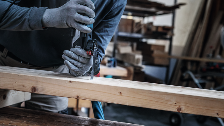 Person wearing safety equipment cuts a wooden beam with a handheld power tool.