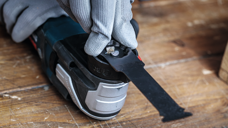 Person wearing safety equipment adjusts a multi-tool blade on a wooden workbench.