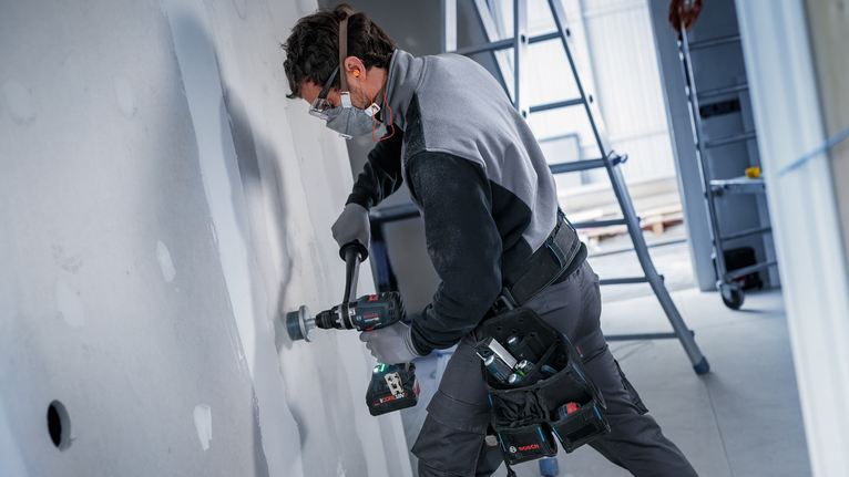 A person wearing safety equipment drills into drywall with a power tool.