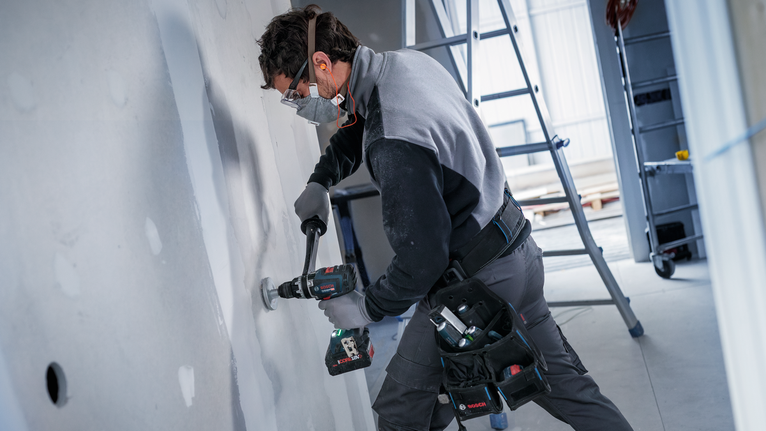 A person wearing safety equipment drills a hole in drywall at a construction site.