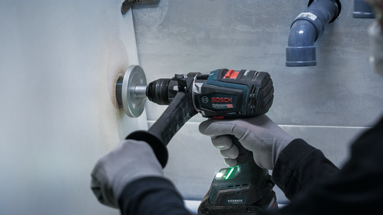 Person wearing safety equipment drills a hole in a wall with a power tool.