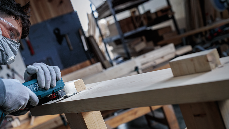 Person wearing safety equipment sands a wooden plank in a workshop.