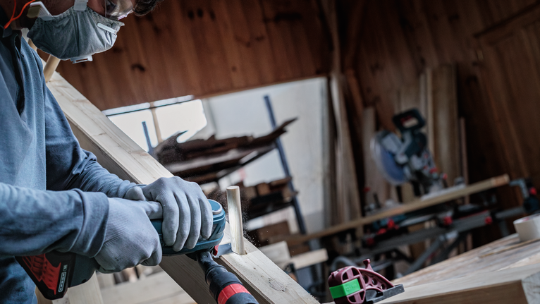 A person wearing safety equipment sands a wooden plank with a power tool.