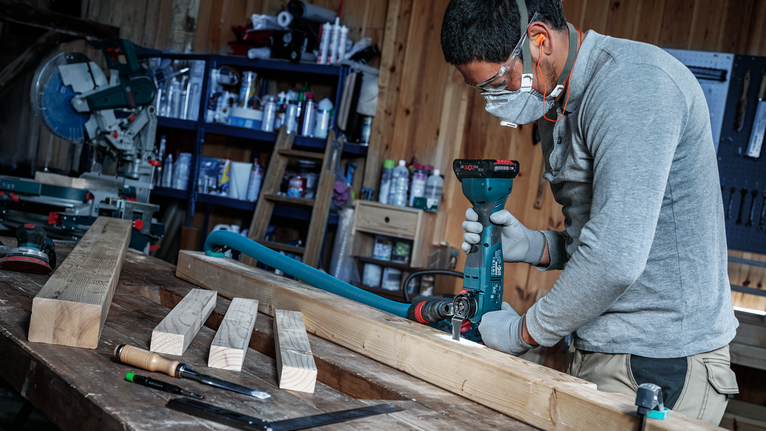 Person wearing safety equipment drills into a wooden beam in a workshop.