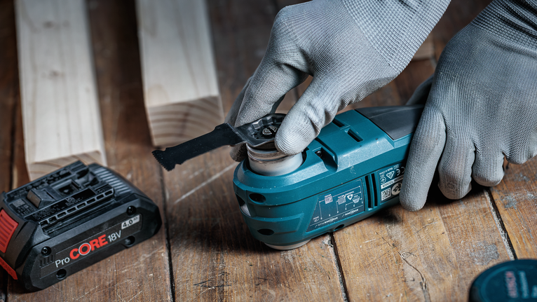 Person wearing safety equipment attaches a blade to a cordless power tool on a wooden surface.