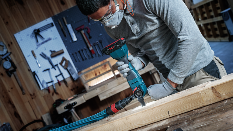 Person wearing safety equipment uses a power tool to cut wood on a workbench.