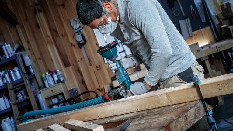 Person wearing safety equipment drills into a wooden beam in a workshop.