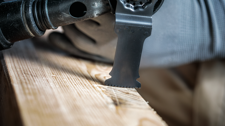 Person wearing safety equipment uses a multitool blade to cut a wooden board.