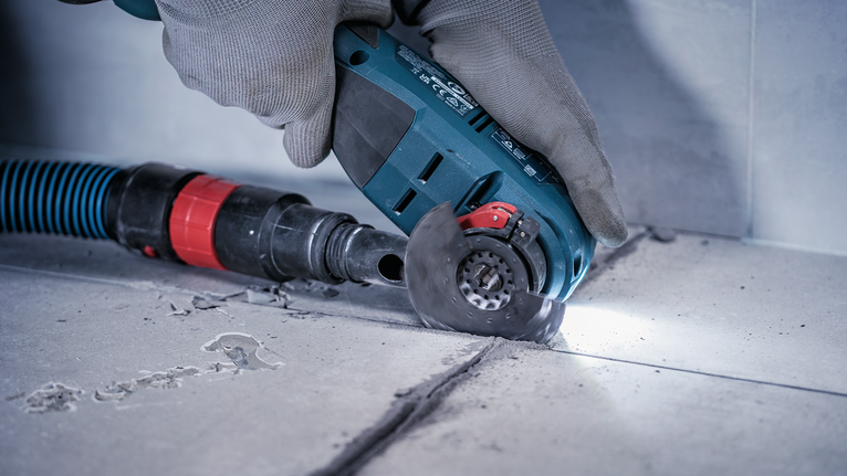 Person wearing safety equipment cuts tile grout with a handheld power tool.