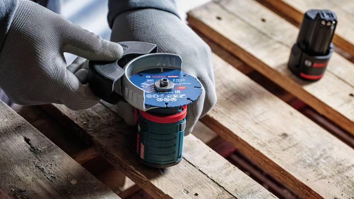 Object #200609214: 2840086_app_web_07 Person wearing safety equipment holds a cordless angle grinder on a wooden pallet.