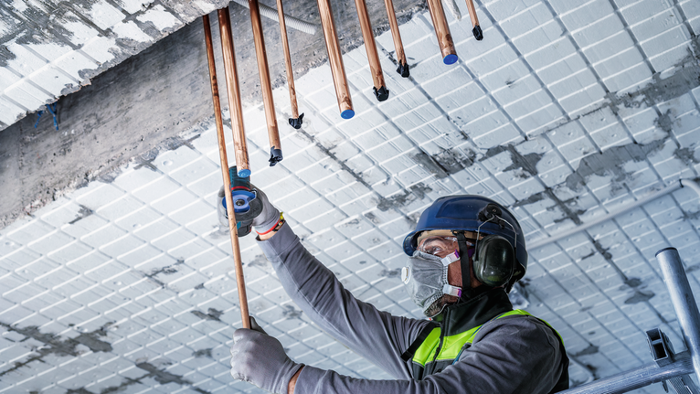 A person wearing safety equipment installs copper pipes into a ceiling.