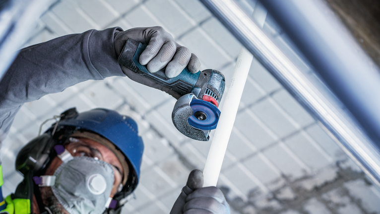 Person wearing safety equipment cuts a white pipe with a compact power tool.