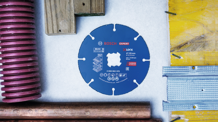 EXPERT Multi Material Carbide Cutting Disc, 125 x 22.23 mm, X-Lock Carbide saw blade placed on a table surrounded by wood, metal, and plastic materials.