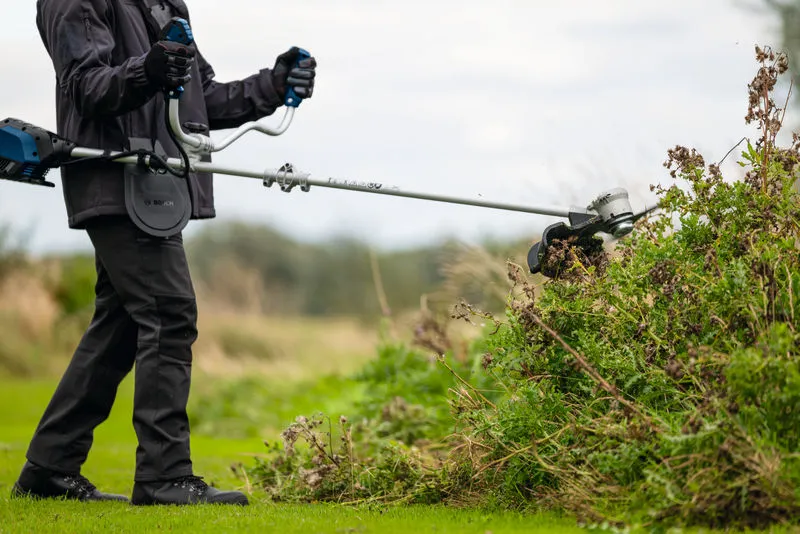 A person wearing safety equipment trims thick brush with a brushcutter outdoors.