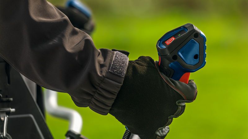 A person wearing safety equipment grips the handle of a brushcutter outdoors.