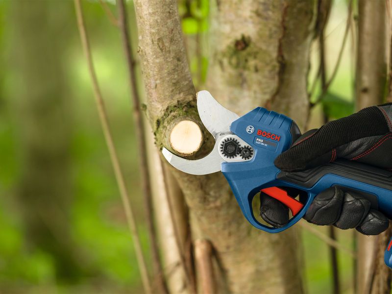 A person wearing safety equipment uses a cordless pruner to cut a tree branch.