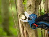 A person wearing safety equipment uses a cordless pruner to cut a tree branch.