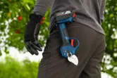A person wearing safety equipment carries a cordless pruner in an orchard.
