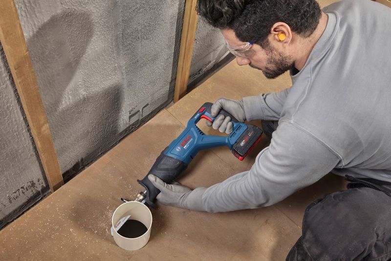 A person wearing safety equipment uses a cordless reciprocating saw to cut a PVC pipe.