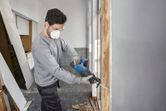 A person wearing safety equipment uses a cordless reciprocating saw to cut a wooden frame.