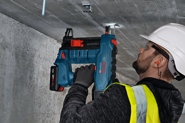 A person wearing safety equipment fastens a metal hanger to a concrete ceiling.