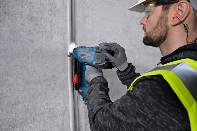 A person wearing safety equipment fastens conduit to a wall using a power tool.
