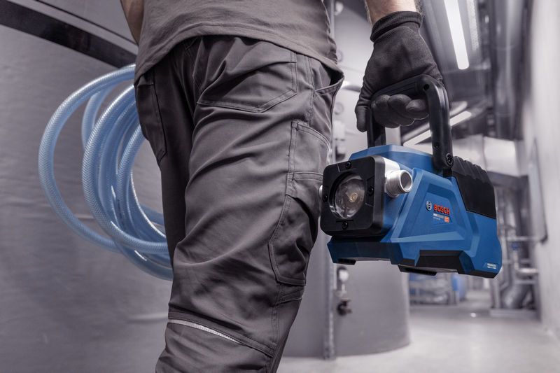 A person wearing safety equipment carries a blue portable vacuum pump in an industrial setting.