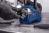 A person wearing safety equipment assembles a blue mechanical device on a metal workbench.