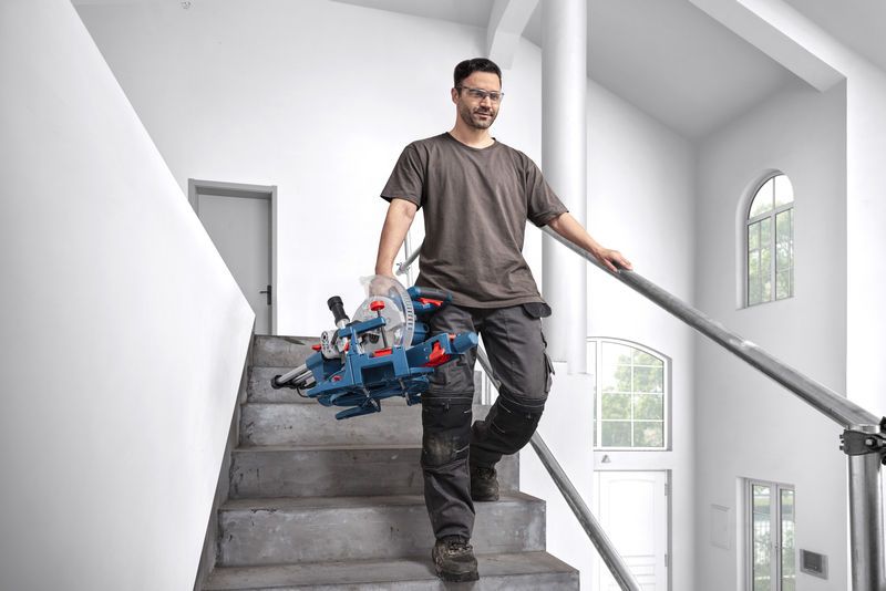 A person wearing safety equipment carries a mitre saw while walking down stairs.