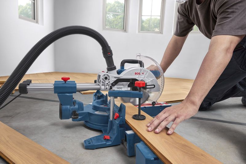 A person wearing safety equipment cuts a wooden plank with a mitre saw.