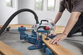 A person wearing safety equipment cuts a wooden plank with a mitre saw.