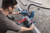 A person wearing safety equipment uses a mitre saw to cut a long metal beam.