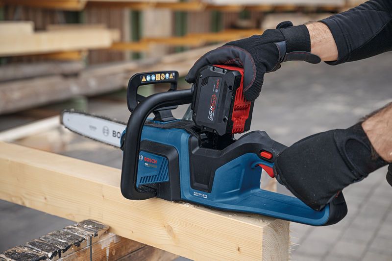 A person wearing safety equipment installs a battery into a cordless chainsaw on a timber beam.