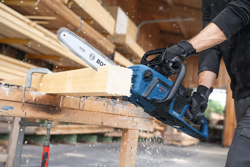 A person wearing safety equipment cuts a wooden beam with a cordless chainsaw.