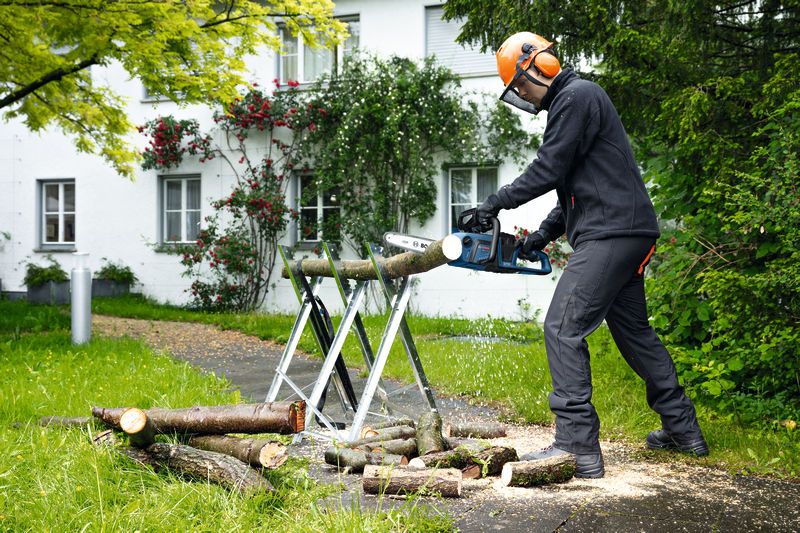 A person wearing safety equipment uses a cordless chainsaw to cut tree branches outdoors.