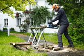 A person wearing safety equipment uses a cordless chainsaw to cut tree branches outdoors.