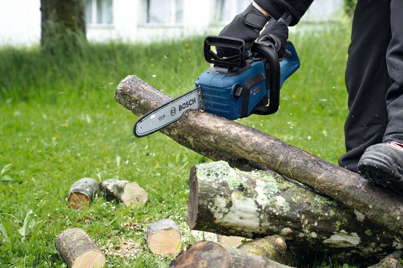 A person wearing safety equipment uses a cordless chainsaw to cut a tree branch.