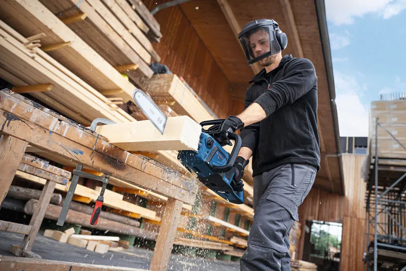 A person wearing safety equipment cuts a wooden beam with a cordless chainsaw.