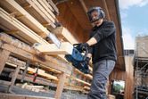 A person wearing safety equipment cuts a wooden beam with a cordless chainsaw.