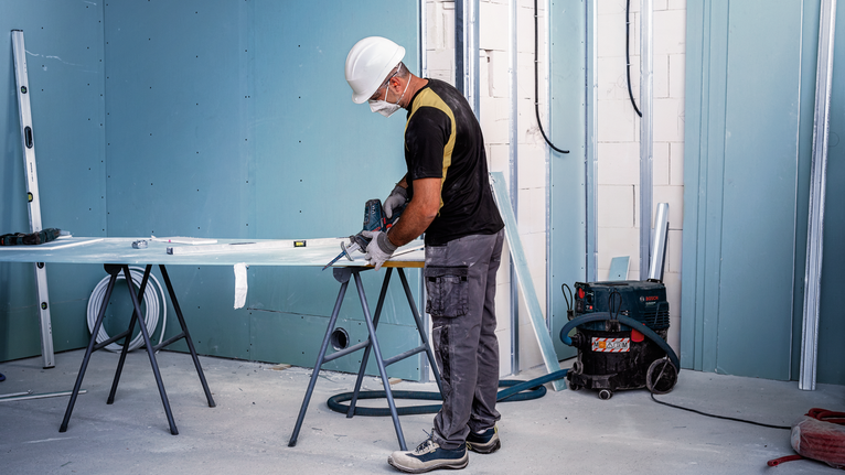 A person wearing safety equipment cuts a board with a power tool in a construction room.